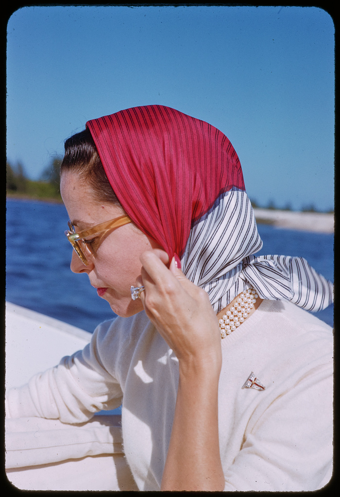 color photograph of gloria guiness in profile on a boat. Gloria is seated and wearing sunglasses with a scarf around her neck and also covering her hair. 