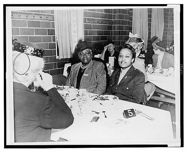Photo of two women looking at the camera while seated at side by side a luncheon table. Other women are seated at other tables in the foreground and background.